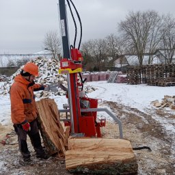 Energia z natury Grzegorz Konecki - Pracownik w kasku obsługuje czerwoną łuparkę do drewna na zewnątrz w zimowy dzień. W tle stos drewna i palety.