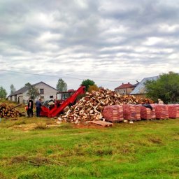 Energia z natury Grzegorz Konecki - Składowanie drewna kominkowego: stos pociętych kawałków, worki na paletach, traktor z podnośnikiem, pracownicy na tle wiejskich zabudowań pod pochmurnym niebem.