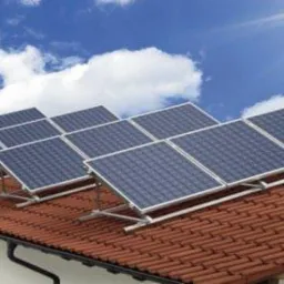 Rząd paneli słonecznych na czerwonym dachu domu, błękitne niebo z promieniami słońca w tle. Widoczne mocowania paneli.