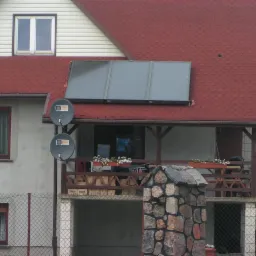 Czerwony dach domu z trzema panelami słonecznymi i dwiema antenami satelitarnymi. Balkon z drewnianą balustradą i kwiatami w doniczkach. Kamienne słupki ogrodzenia.