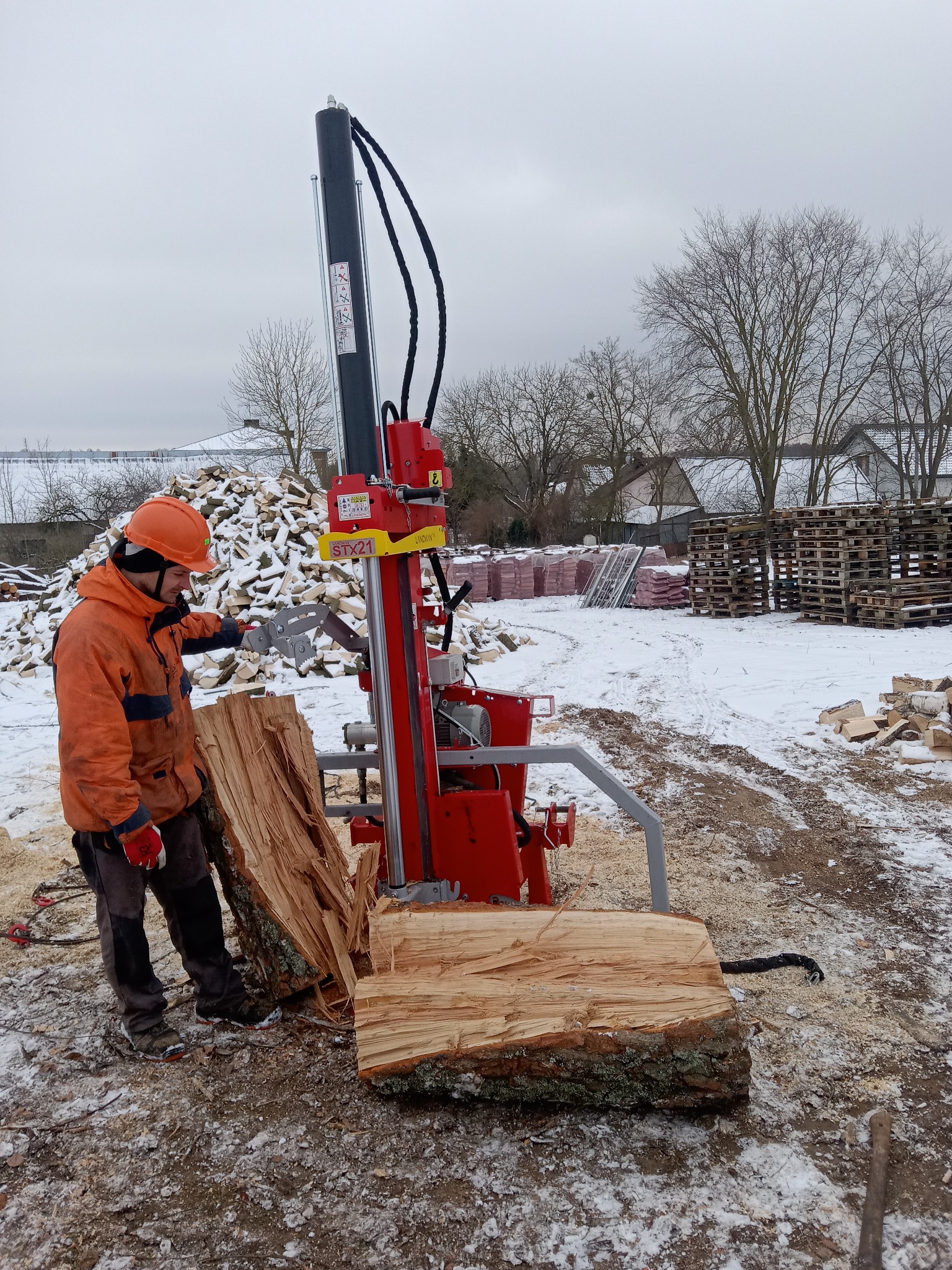 Pracownik w kasku obsługuje czerwoną łuparkę do drewna na zewnątrz w zimowy dzień. W tle stos drewna i palety.