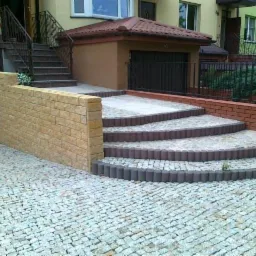 Wykończone schody wejściowe z kostki brukowej prowadzące do domu. Widoczny murek z bloczków i podjazd z kostki granitowej. Estetyczne i funkcjonalne rozwiązanie.