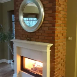 Kominek z cegły z białym obramowaniem i paleniskiem. Nad nim okrągłe lustro i dekoracyjna figurka słonia. Widoczne płomienie w palenisku.