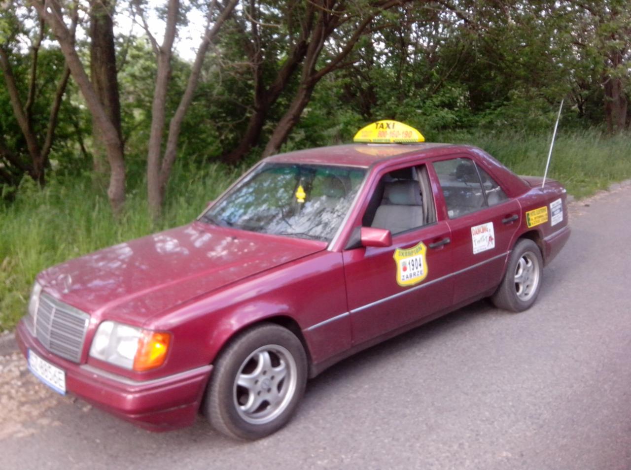 Czerwony Mercedes Taxi z żółtym szyldem na dachu, z naklejkami na karoserii, zaparkowany w Zabrzu, na tle zieleni. Widok z boku, lekko z góry.