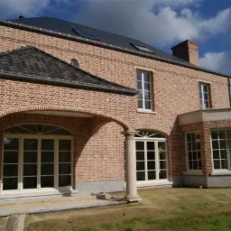 Murowany dom z czerwonej cegły z dużymi oknami z białymi ramami i przeszkleniami, widoczne również okna dachowe i kolumna podtrzymująca łuk