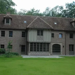 Duży, dwupiętrowy dom z cegły z wieloma oknami i drzwiami tarasowymi, kryty brązową dachówką, otoczony zielonym trawnikiem i drzewami.