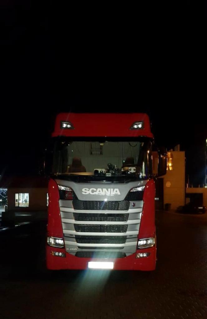 Czerwono-szara ciężarówka Scania sfotografowana z przodu w nocy, z widocznymi światłami i odblaskami, na tle ciemnego otoczenia z budynkami w oddali.