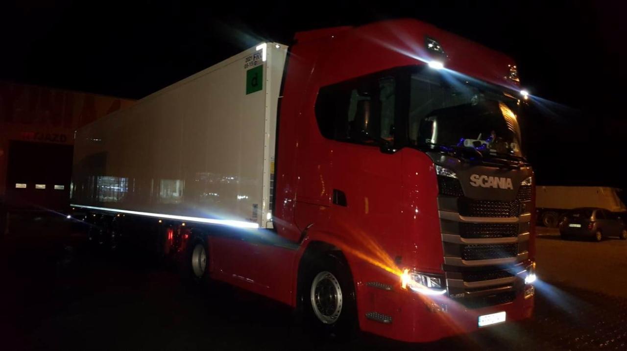 Czerwona ciężarówka Scania z białą naczepą chłodniczą, oświetlona reflektorami, stoi na parkingu w nocy.