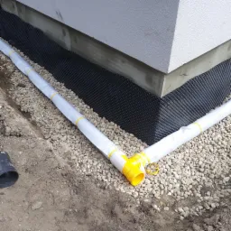 Czarna folia kubełkowa przy fundamencie budynku, zabezpieczająca przed wilgocią, z ułożonym systemem rur drenarskich w białej geowłókninie, obsypanych drobnymi kamieniami.