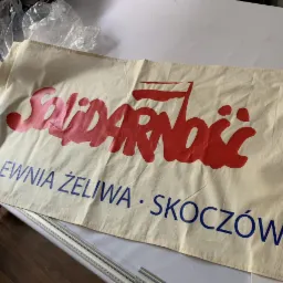 Beżowy baner z czerwonym napisem 'Solidarność' i niebieskim tekstem 'Odlewnia Żeliwa Skoczów' leżący na stole.