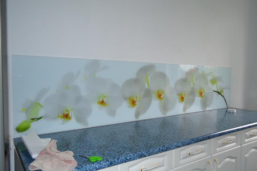 Szklany panel ścienny z motywem białych orchidei nad blatem kuchennym, widoczne narzędzia montażowe i elementy wykończeniowe.
