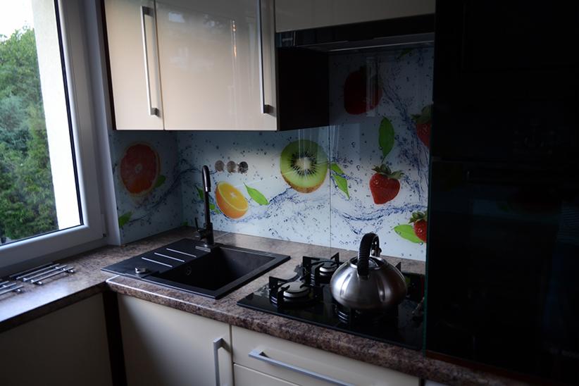 Panel szklany w kuchni z motywem owoców (pomarańcza, kiwi, truskawka, grejpfrut) i efektem rozpryskującej się wody, zamontowany nad zlewem i płytą grzewczą.