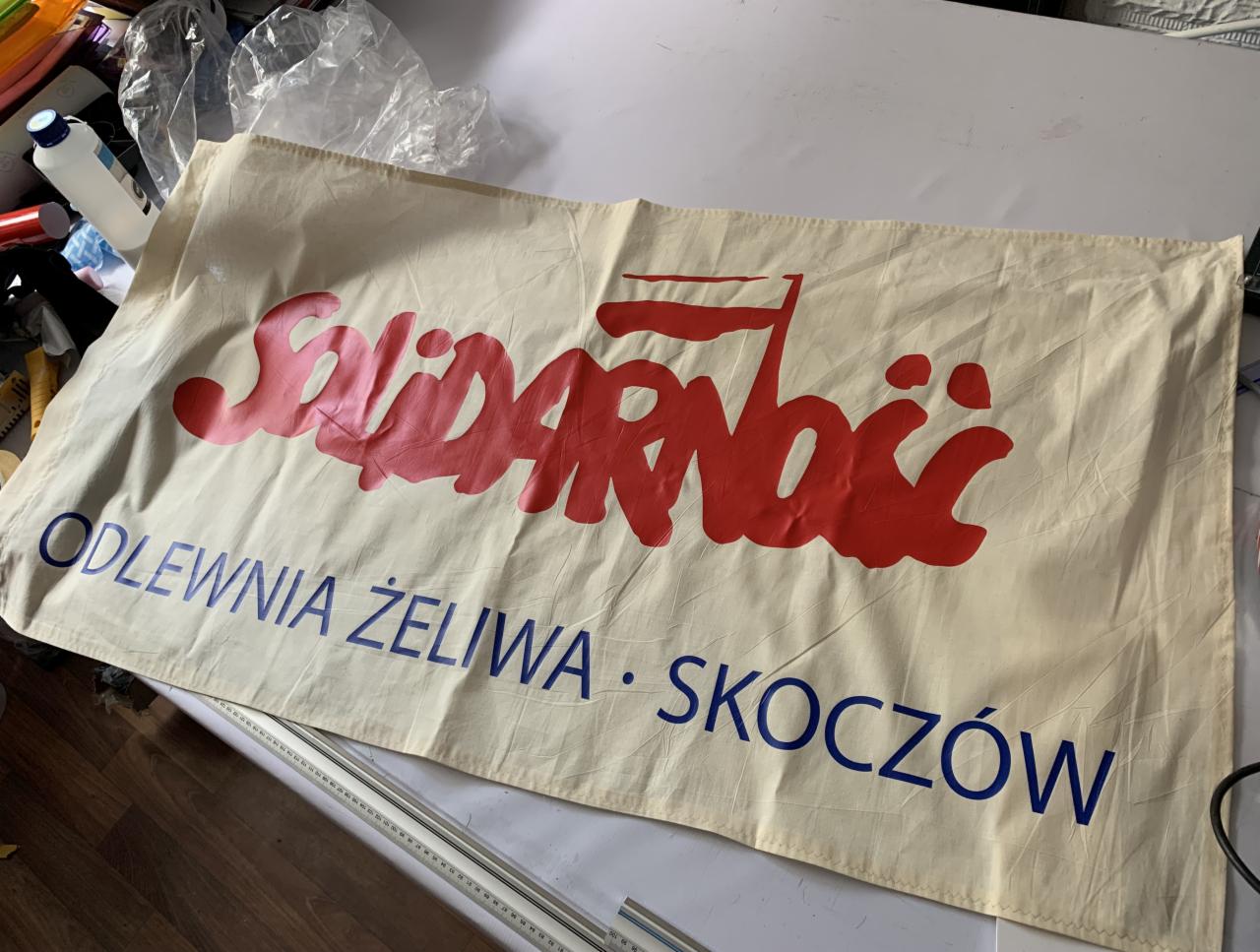 Beżowy baner z czerwonym napisem 'Solidarność' i niebieskim tekstem 'Odlewnia Żeliwa Skoczów' leżący na stole.