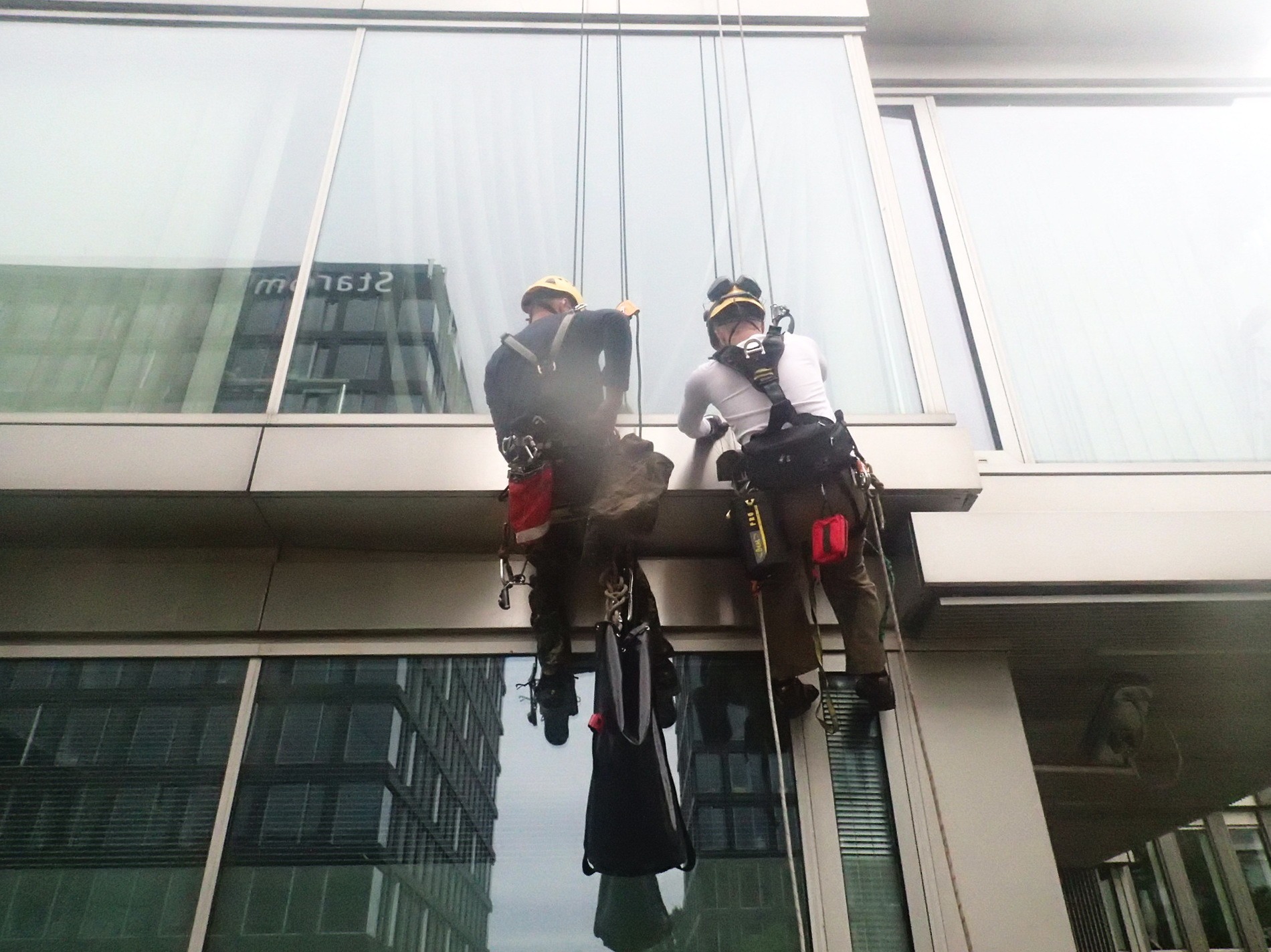 Dwóch pracowników wysokościowych, zabezpieczonych linami, podczas pracy na szklanej elewacji biurowca; widoczne narzędzia i odblaskowe elementy odzieży.