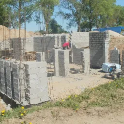 Budowa domu z betonu komórkowego: fundamenty, ściany w trakcie wznoszenia, widoczna praca murarza w czerwonej koszulce na tle zieleni.