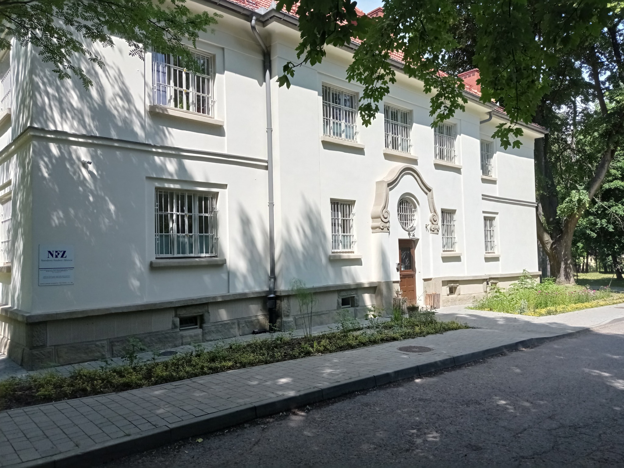 Biały, dwupiętrowy budynek Szpitala im. Babińskiego w Krakowie z widoczną tablicą NFZ, okna zabezpieczone kratami, otoczony zielenią i chodnikiem z kostki brukowej.