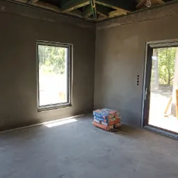 Surowe wnętrze w trakcie prac wykończeniowych: ściany pokryte świeżo nałożonym tynkiem, widoczne worki z zaprawą murarską ułożone w rogu pomieszczenia, surowy beton na podłodze, okno z widokiem...