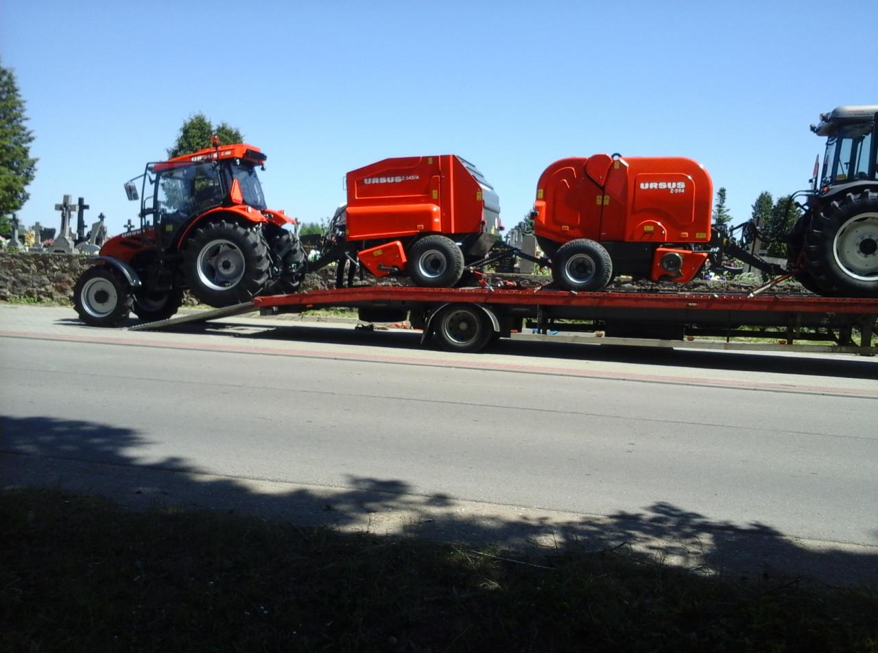 Laweta przewozi dwa czerwone traktory Ursus i prasę rolniczą na tle cmentarza, ukazując transport ciężarowy sprzętu rolniczego w słoneczny dzień.