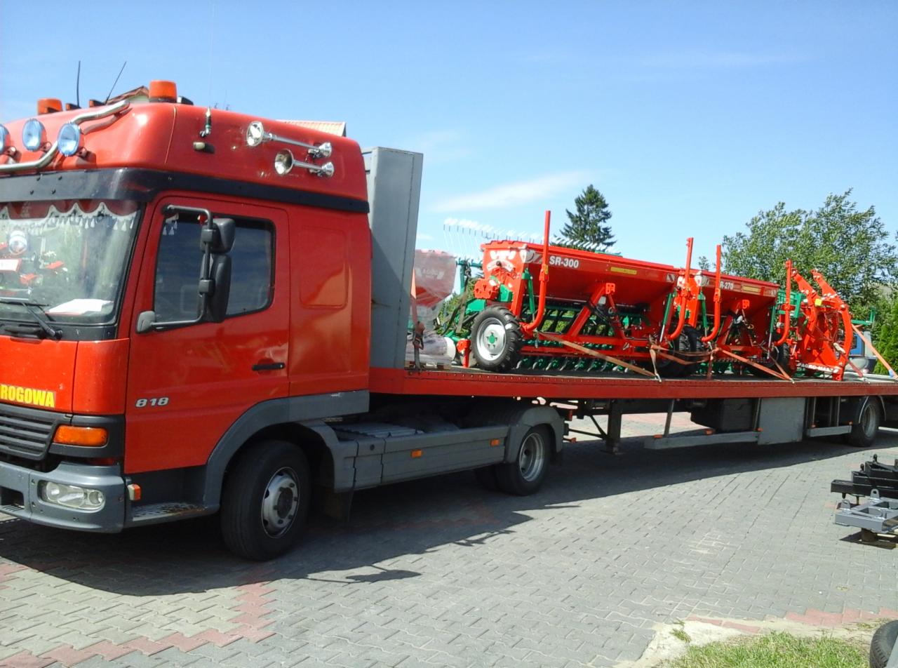 Czerwona laweta przewozi dwie czerwone maszyny rolnicze (siewniki) marki SR-300 na platformie. Widoczne drzewa i błękitne niebo w tle. Transport specjalistyczny.
