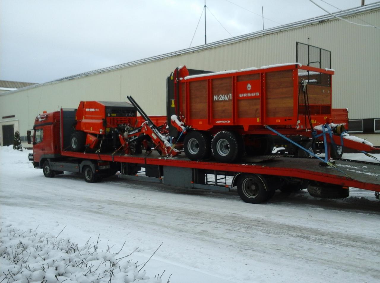 Laweta przewozi sprzęt rolniczy marki Ursus w zimowych warunkach. Na platformie widoczny ciągnik i przyczepa. Tło stanowi hala przemysłowa i śnieżny krajobraz.