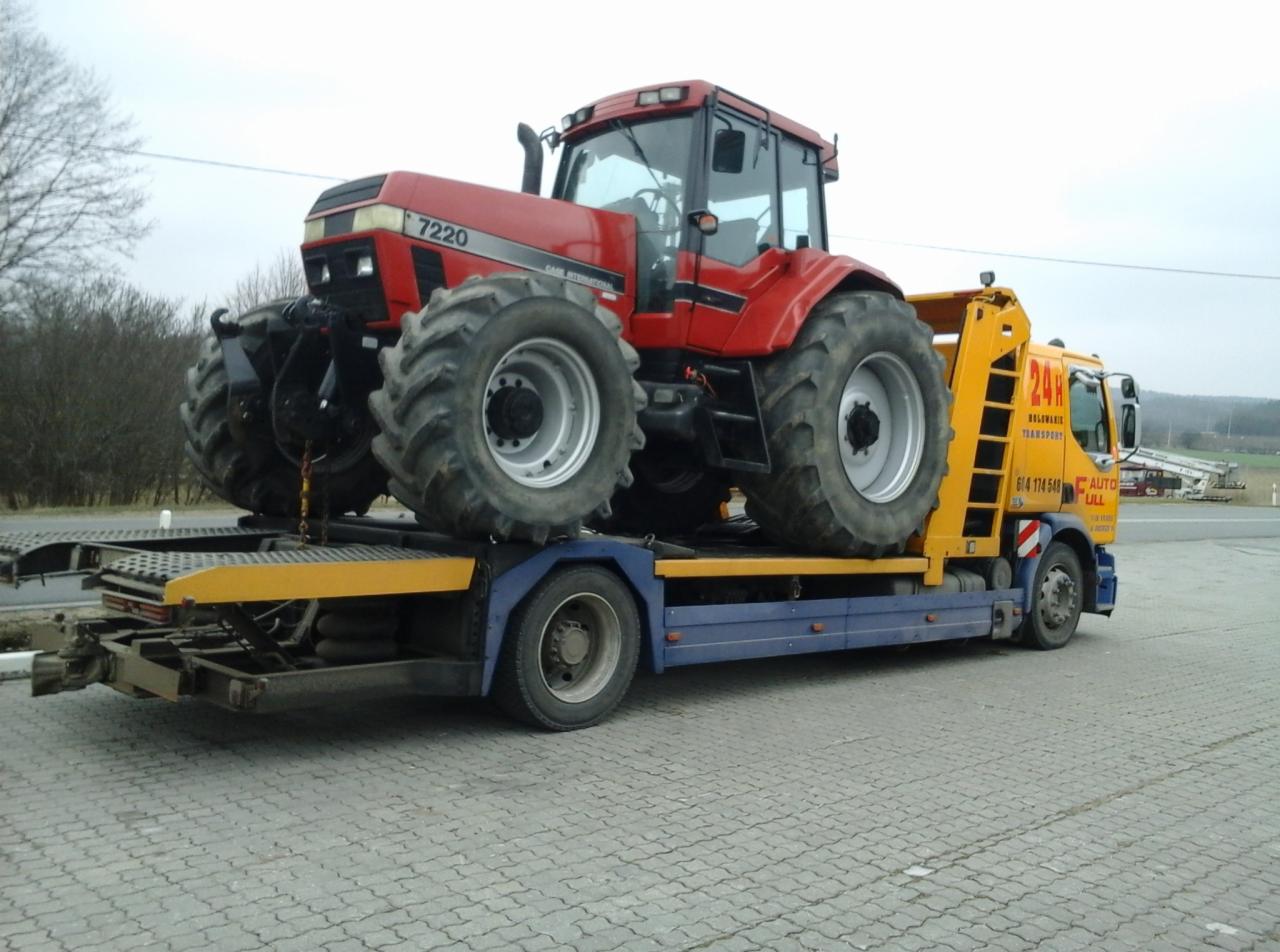 Czerwony ciągnik Case 7220 na żółto-niebieskiej lawecie. Transport maszyny rolniczej na tle krajobrazu polnego. Widoczne oznaczenia firmy transportowej.