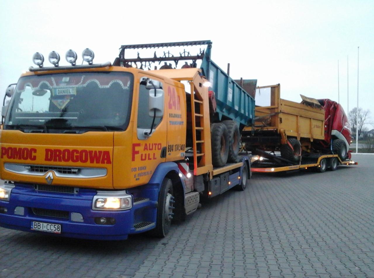 Żółto-niebieska laweta z napisem 'Pomoc Drogowa' przewozi dwie przyczepy rolnicze na platformie. Widok z boku na szarym tle.