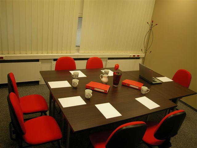 Sala konferencyjna z ciemnym stołem, czerwonymi krzesłami, laptopem, filiżankami, dzbankiem, segregatorami i kartkami papieru, oświetlona sztucznym światłem z roletami w tle.