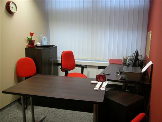 Biuro z dwoma czerwonymi krzesłami, biurkiem z ciemnego drewna, komputerem, drukarką i zegarem na ścianie, oświetlone przez rolety pionowe.