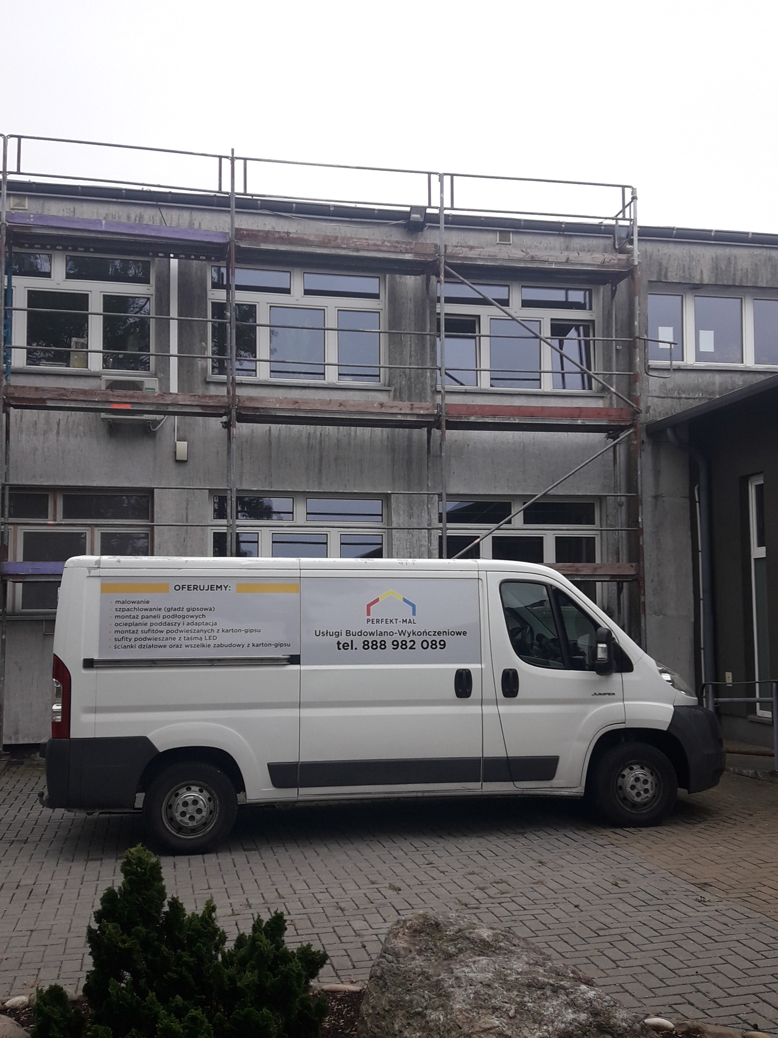 Budynek w trakcie remontu, widoczne rusztowania i samochód dostawczy z reklamą usług budowlano-wykończeniowych.
