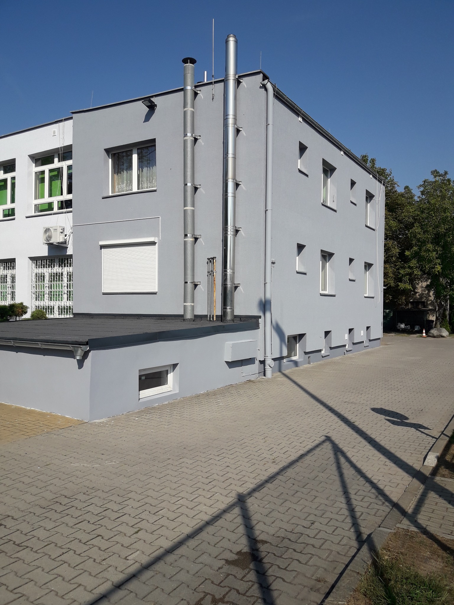 Dwukondygnacyjny budynek pomalowany na szaro z metalowymi kominami na zewnątrz, widoczne okna z białymi ramami i roletą, chodnik z kostki brukowej.