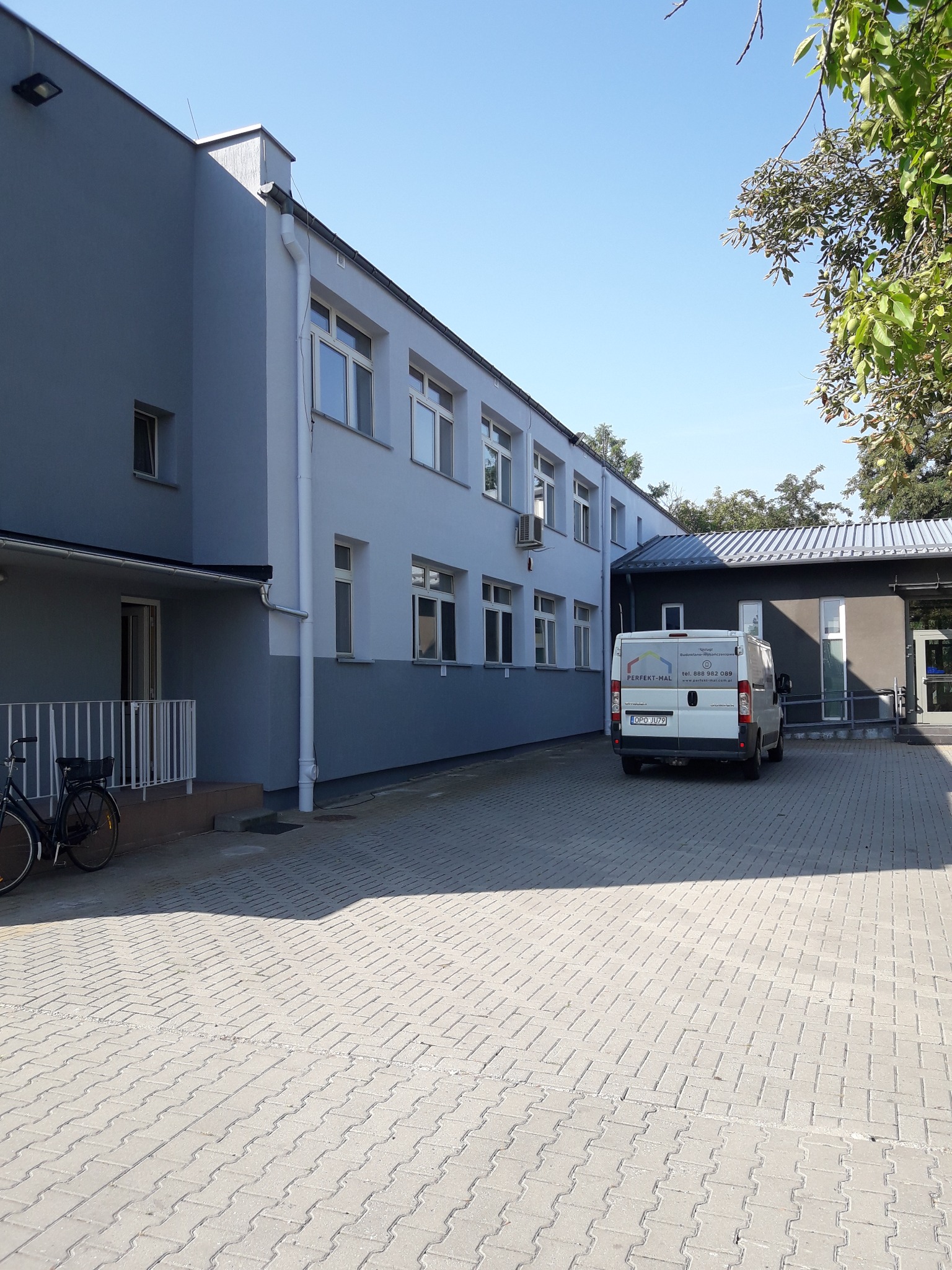 Odnowiony budynek z dwukolorową elewacją w odcieniach szarości, z zaparkowanym białym vanem firmy remontowej i rowerem opartym o barierkę.