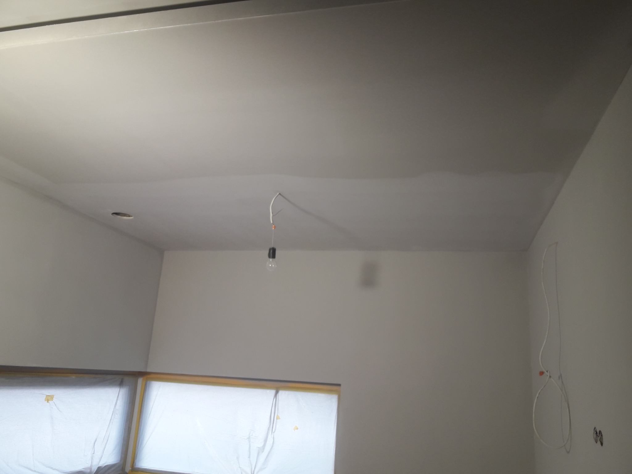 Świeżo pomalowany sufit w pokoju, widoczne zabezpieczone folią okna, zwisająca żarówka i ślady po szpachlowaniu.