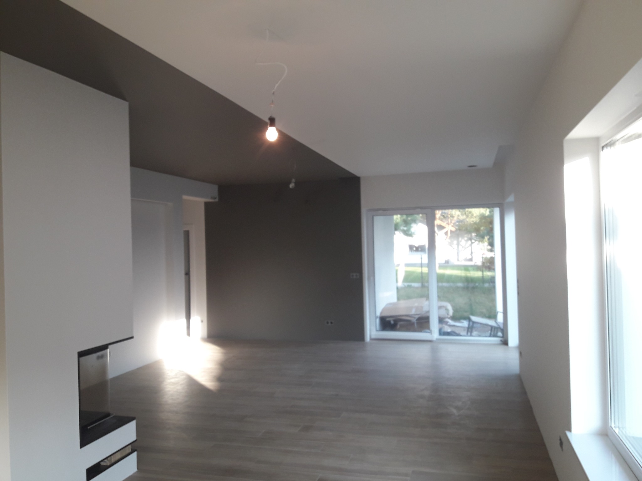 Surowe wnętrze salonu po częściowym pomalowaniu, z szarą ścianą i sufitem kontrastującymi z białymi ścianami, widoczną instalacją elektryczną i panelami podłogowymi.