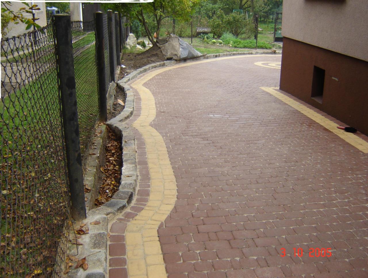 Ułożona z czerwonej kostki brukowej, zakrzywiona ścieżka z żółtymi akcentami, oddzielona od trawnika niskim murkiem z kamienia i metalowym ogrodzeniem.