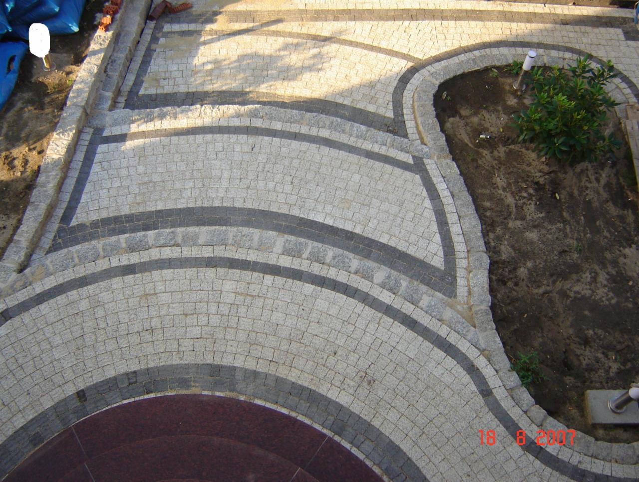 Ułożona z kostki brukowej ścieżka ogrodowa z elementami dekoracyjnymi w postaci ciemniejszych pasów i obrzeży, widok z góry na fragment ogrodu z roślinami.