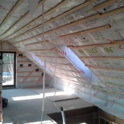 Poddasze w trakcie ocieplania pianką, widoczne krokwie, folia paroizolacyjna z nadrukiem 'Intello', okno dachowe i drabina aluminiowa.