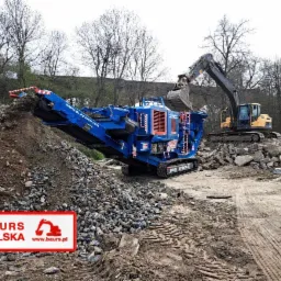 Żółta koparka ładuje gruz do niebieskiego kruszera mobilnego 900x600 METROTRACK. W tle drzewa, a w lewym dolnym rogu logo firmy Baur's Polska.