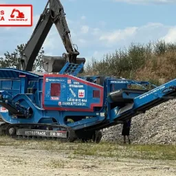 Mobilny kruszacz szczękowy Terex Pegson Metrotrak 900x600 w kolorze niebieskim z logo firmy Baur's Polska, na tle zieleni i gruzu.