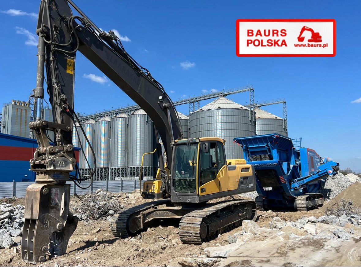 Żółta koparka z hydraulicznym nożem kruszącym i niebieski kruszarka Rex Pegson na placu budowy, w tle silosy i logo firmy Baur's Polska.