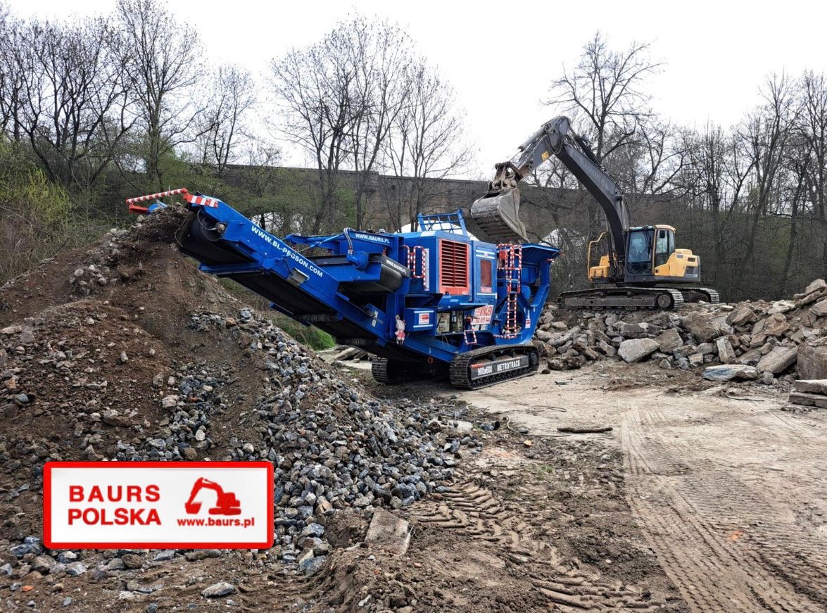 Żółta koparka ładuje gruz do niebieskiego kruszera mobilnego 900x600 METROTRACK. W tle drzewa, a w lewym dolnym rogu logo firmy Baur's Polska.