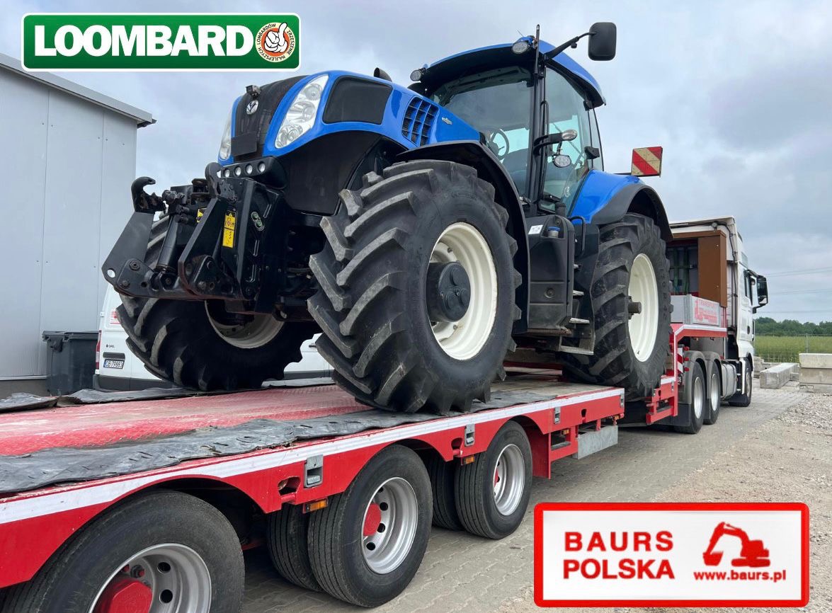 Niebieski traktor na czerwonej platformie transportowej, widok z boku, logo Loombard w rogu. Widoczne koła ciągnika i naczepy. Transport maszyny rolniczej.
