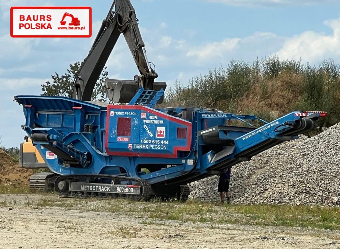 Mobilny kruszacz szczękowy Terex Pegson Metrotrak 900x600 w kolorze niebieskim z logo firmy Baur's Polska, na tle zieleni i gruzu.