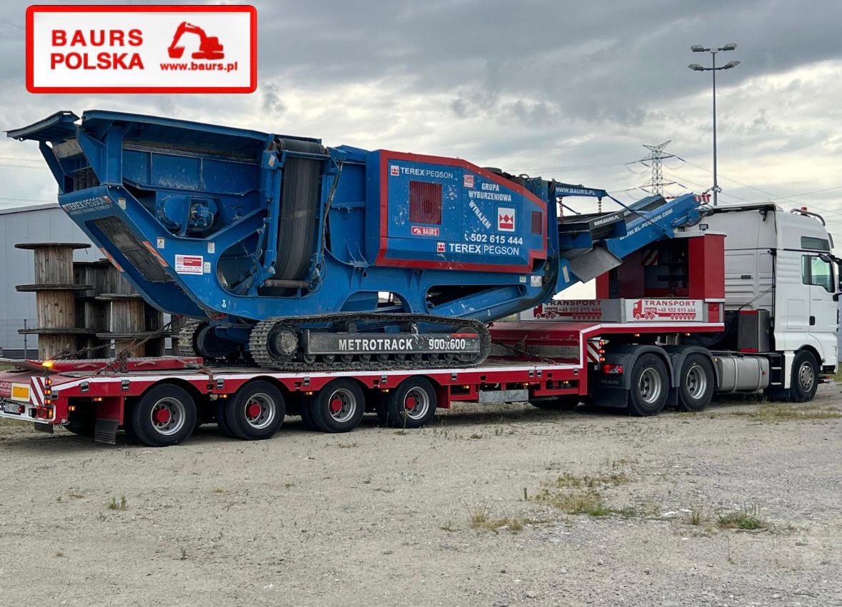 Niebieska kruszarka Terex Pegson Metrotrak 900x600 na czerwonej platformie niskopodwoziowej, przewożona przez białą ciężarówkę MAN. Widoczne logo firmy Baurs Polska.