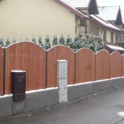 Nowoczesne ogrodzenie drewniane z półokrągłym wykończeniem, otulone lekkim śniegiem, na tle zabudowy szeregowej i zimozielonych drzew.