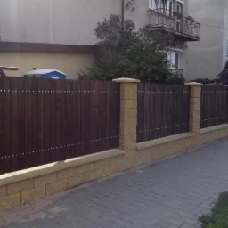 Nowoczesne ogrodzenie z ciemnego drewna z elementami z piaskowca, widoczne detale mocowania na tle budynku mieszkalnego.