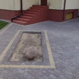 Ułożona szara kostka brukowa typu nostalit firmy Jadar z obramowaniem w kolorze piaskowym, tworząca ścieżkę i podjazd przed budynkiem z ceglanym cokołem i schodami; w centralnym punkcie wyznaczony...