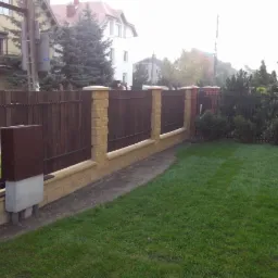 Nowoczesne ogrodzenie z drewnianymi panelami i kamiennymi słupkami, skrzynka na listy przy wejściu, świeżo założony trawnik z rolki, znak drogowy '40' w tle.