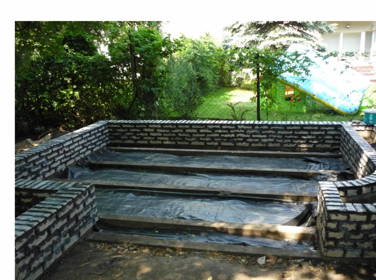 Fundament pod taras z cegły klinkierowej w trakcie budowy, widoczne legary i folia izolacyjna, w tle trawnik i zjeżdżalnia dmuchana.