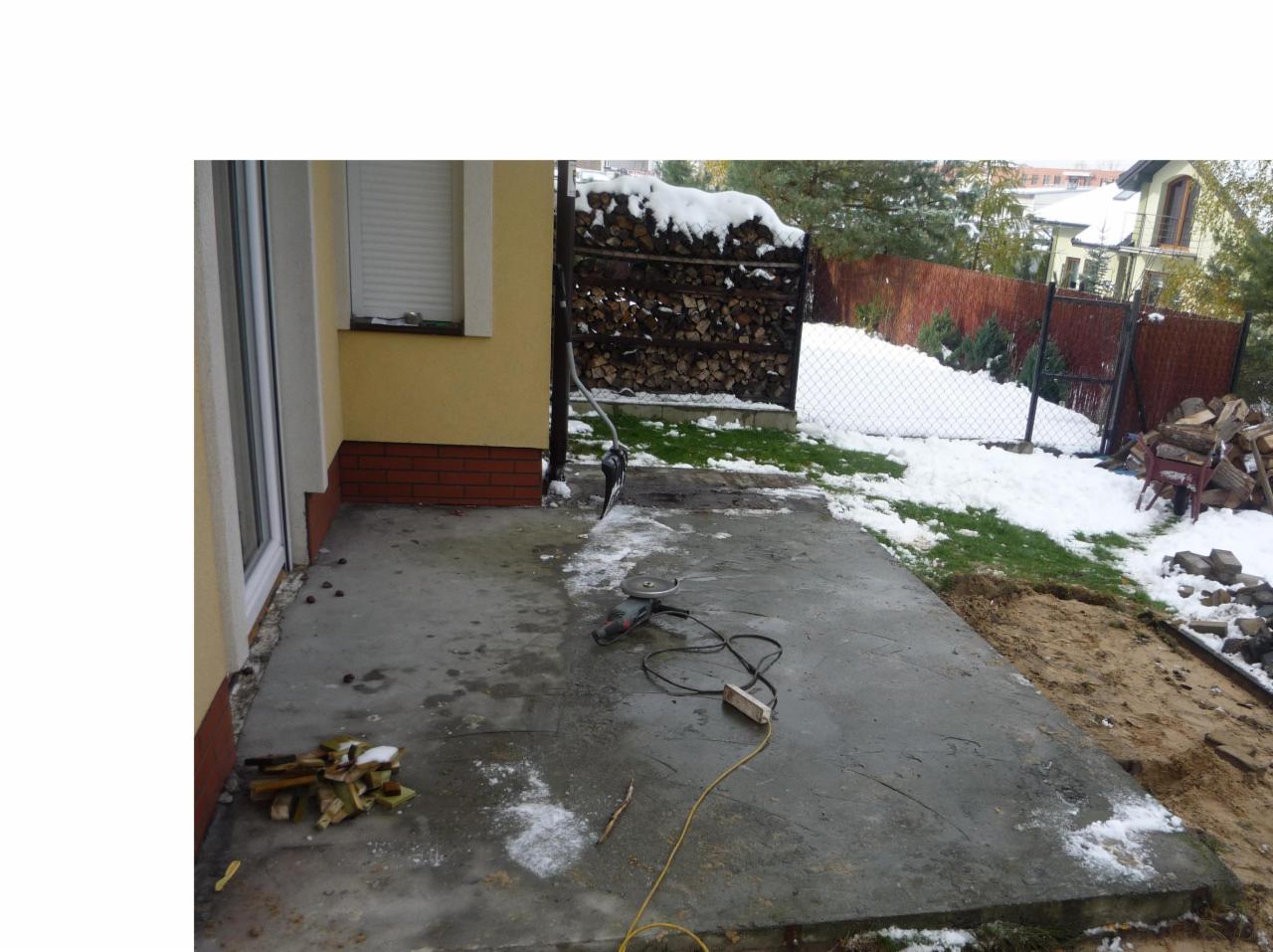 Przygotowanie podłoża pod taras betonowy: widoczny betonowy fundament, szlifierka kątowa, kable, fragmenty drewna i śnieg. W tle ściana budynku z oknem i stosem drewna opałowego, a dalej ogrodzenie...