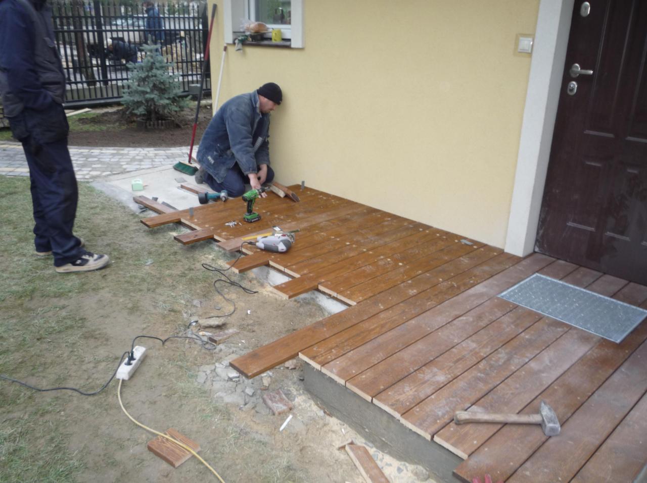 Prace montażowe przy budowie tarasu z desek modrzewiowych przy wejściu do domu; widoczny pracownik w trakcie wkręcania desek, narzędzia pracy (wkrętarka, poziomica), częściowo ułożona podłoga tarasu.